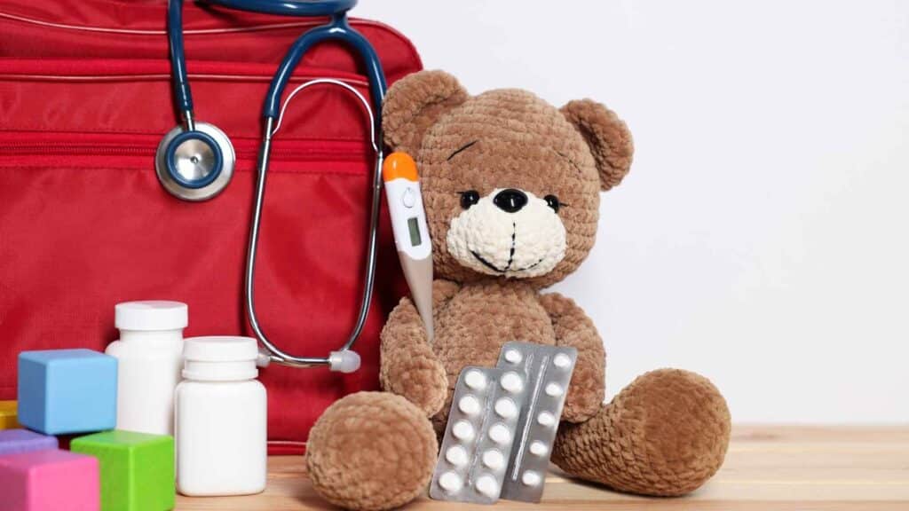 Farmacia da viaggio per bambini: medicinali, termometro e kit medico accanto a un peluche su un tavolo di legno.