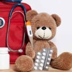 Farmacia da viaggio per bambini: medicinali, termometro e kit medico accanto a un peluche su un tavolo di legno.