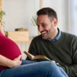 Un uomo sorridente legge un libro vicino al pancione della compagna incinta, illustrando come il feto possa sentire le voci e stabilire una relazione prima della nascita.