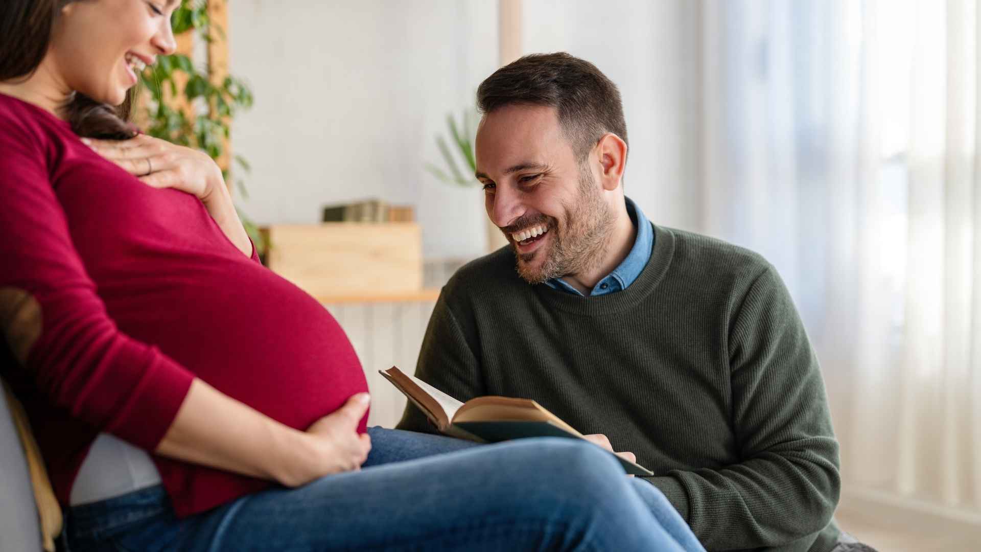 Un uomo sorridente legge un libro vicino al pancione della compagna incinta, illustrando come il feto possa sentire le voci e stabilire una relazione prima della nascita.