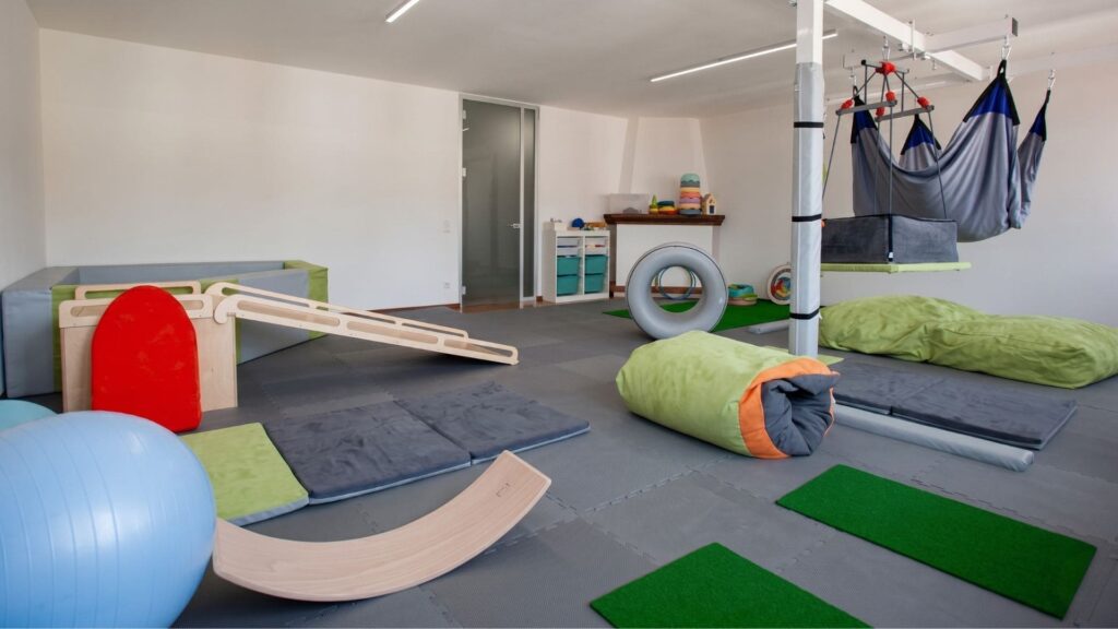 Panoramica di una sala di ergoterapia e integrazione sensoriale vista dall'interno di una vasca con palline trasparenti. La stanza presenta grandi finestre luminose, amache terapeutiche sospese a una struttura, grandi cuscini morbidi verdi (crash pad) e attrezzature per stimoli propriocettivi e vestibolari.