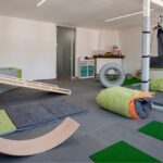 Panoramica di una sala di ergoterapia e integrazione sensoriale vista dall'interno di una vasca con palline trasparenti. La stanza presenta grandi finestre luminose, amache terapeutiche sospese a una struttura, grandi cuscini morbidi verdi (crash pad) e attrezzature per stimoli propriocettivi e vestibolari.