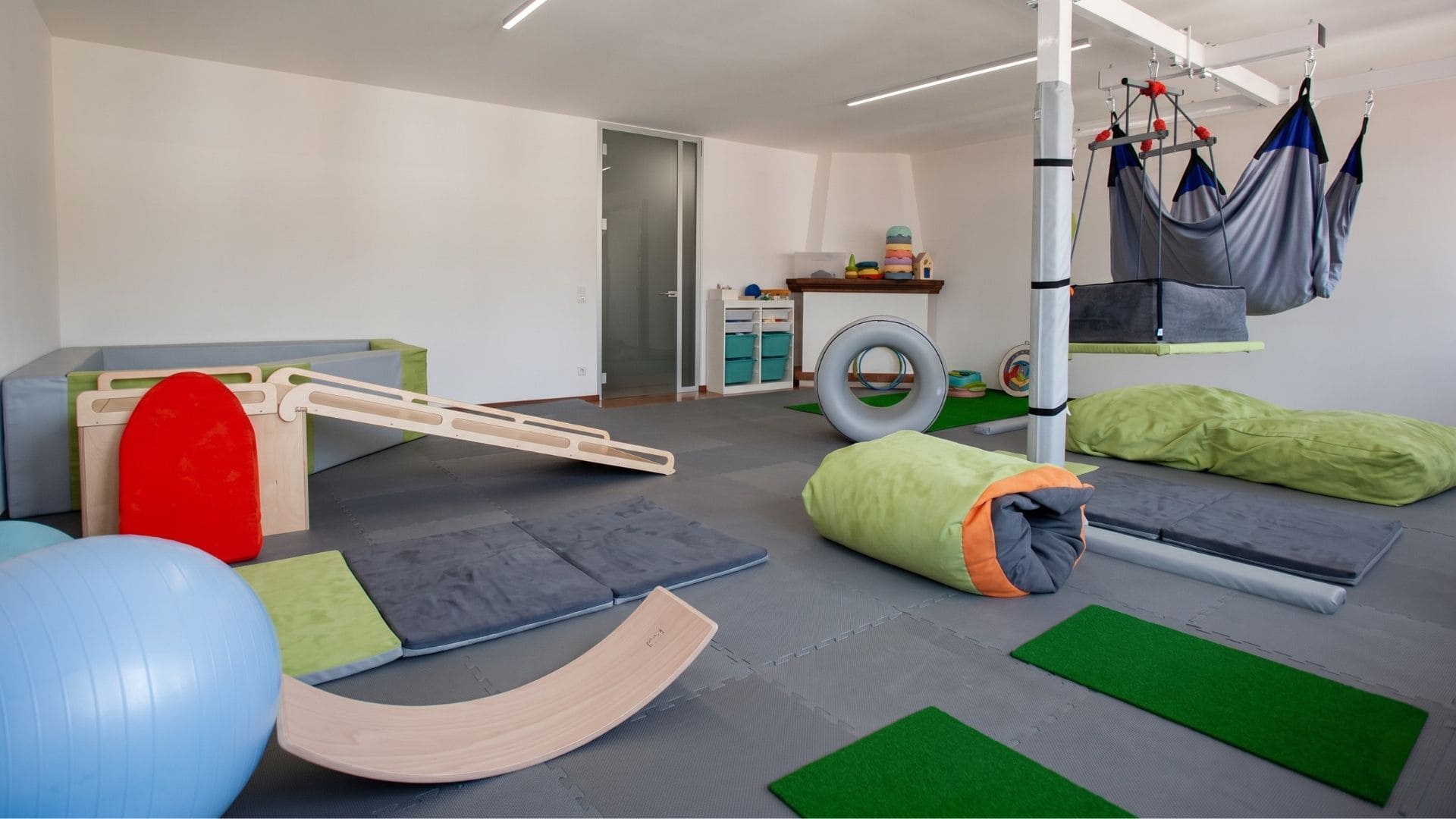 Panoramica di una sala di ergoterapia e integrazione sensoriale vista dall'interno di una vasca con palline trasparenti. La stanza presenta grandi finestre luminose, amache terapeutiche sospese a una struttura, grandi cuscini morbidi verdi (crash pad) e attrezzature per stimoli propriocettivi e vestibolari.