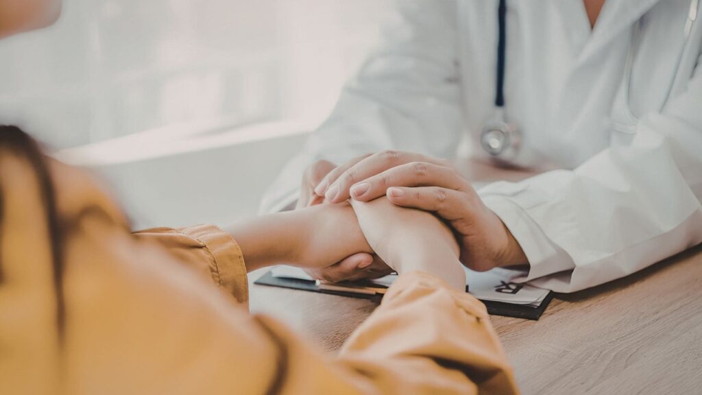 Primo piano di un medico in camice bianco che stringe calorosamente ed empaticamente le mani di una paziente seduta di fronte a lui durante un colloquio in studio.