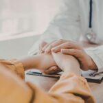 Primo piano di un medico in camice bianco che stringe calorosamente ed empaticamente le mani di una paziente seduta di fronte a lui durante un colloquio in studio.