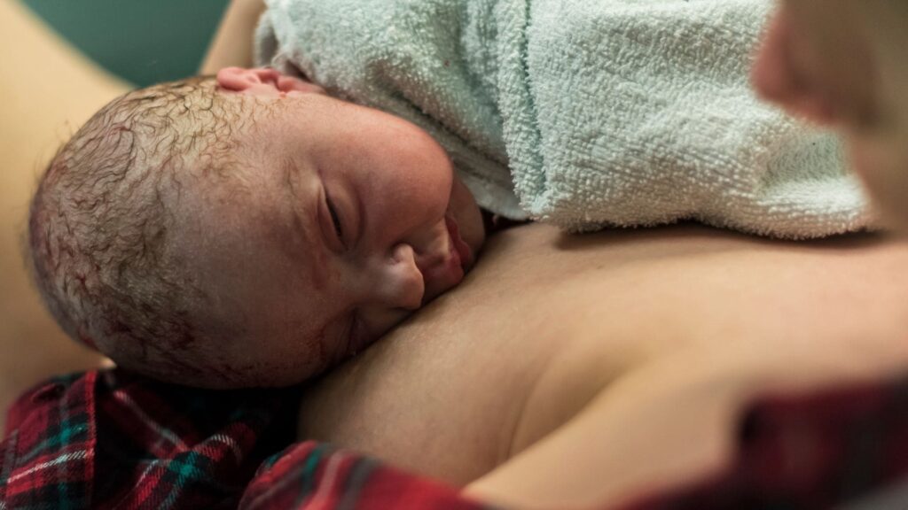 Un neonato che si appoggia delicatamente al petto del genitore, coperto da un asciugamano di spugna color acqua, che mostra i primi istanti di adattamento del neonato alla nascita attraverso il contatto pelle a pelle.