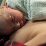 Un neonato che si appoggia delicatamente al petto del genitore, coperto da un asciugamano di spugna color acqua, che mostra i primi istanti di adattamento del neonato alla nascita attraverso il contatto pelle a pelle.